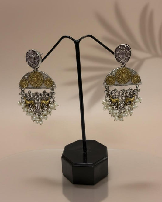 ChicShukra brass oxidised silver-gold jhumka earrings with pearl drops and floral motifs on a black display stand.