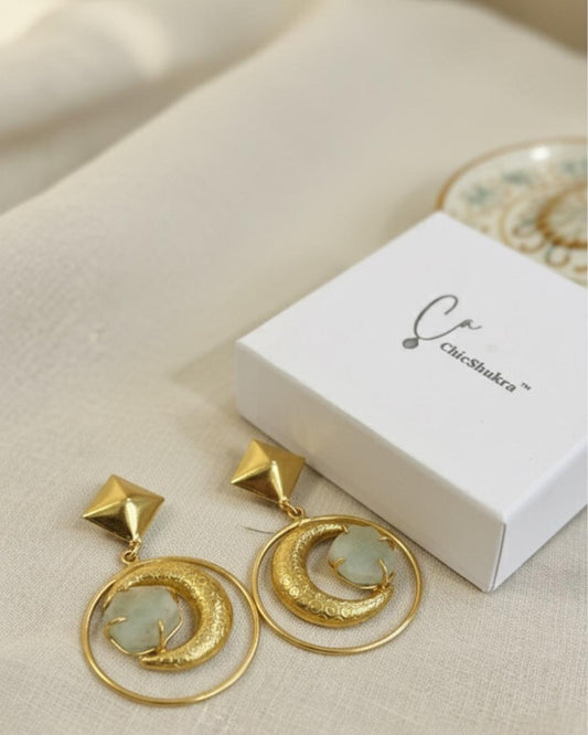 Gold‑tone brass crescent moon earrings with pale stone and pyramid studs beside a white ChicShukra branded box on fabric.
