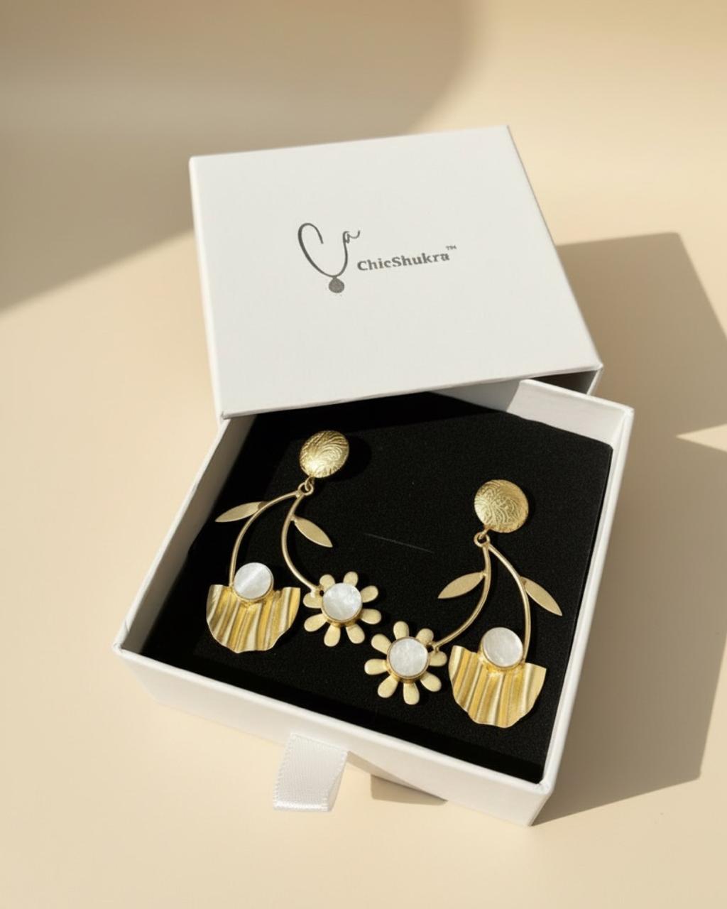 Gold‑toned ChicShukra brass floral earrings with white stone accents displayed on a black earring stand.