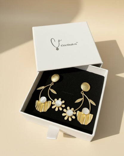 Gold‑toned ChicShukra brass floral earrings with white stone accents displayed on a black earring stand.