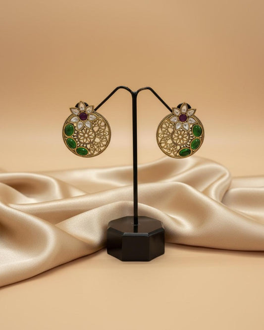 Gold earrings with green and purple stones on a black stand against a beige fabric background