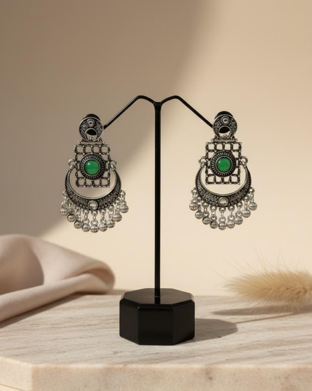 ChicShukra silver‑tone imitation jewellery earrings with green gemstone and dangling beads displayed on a black stand against a beige backdrop.