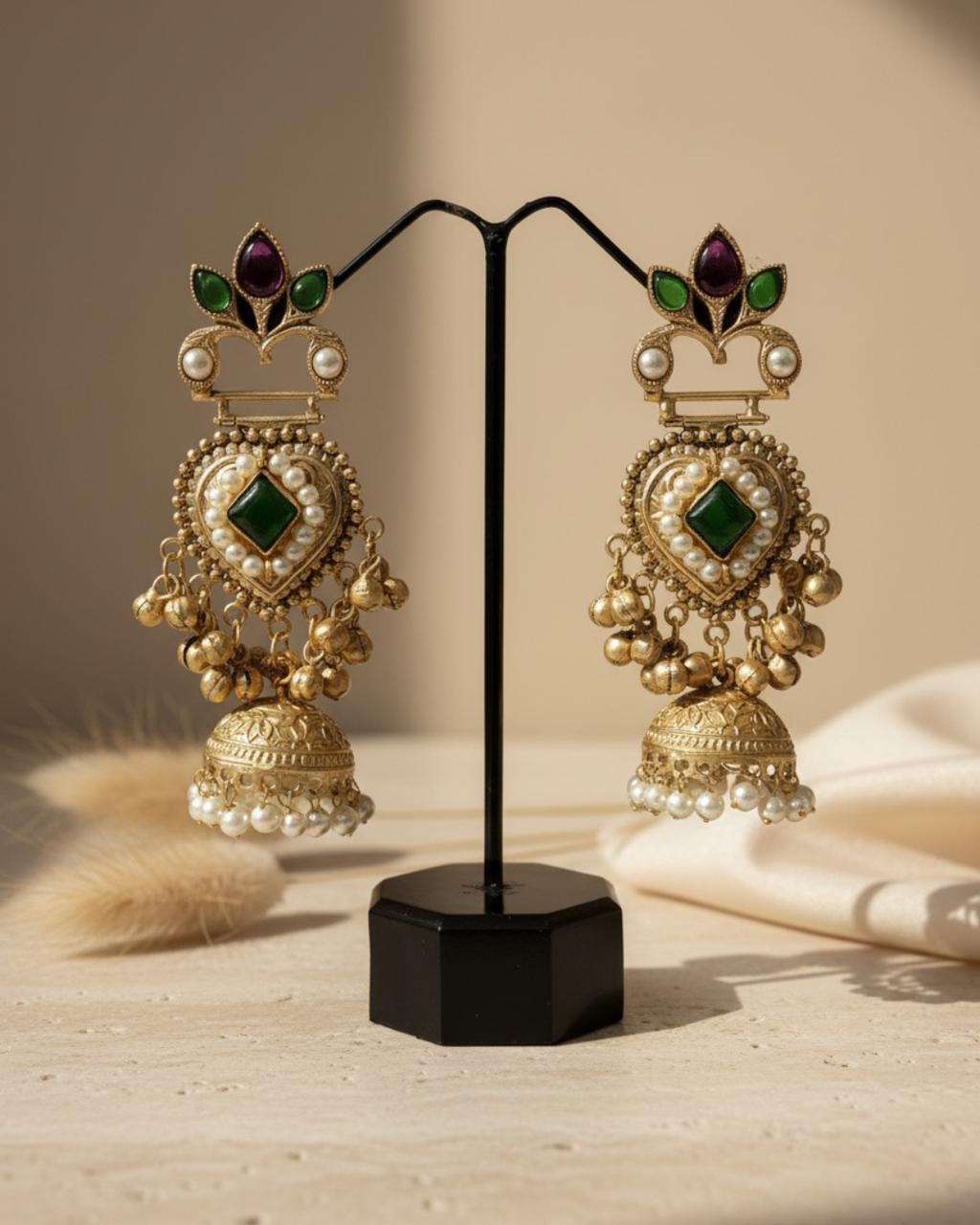 ChicShukra gold‑tone jhumka earrings with purple gem crown, green leaf accents, pearl‑lined heart center, and pearl‑trimmed jhumka bells on a display stand.