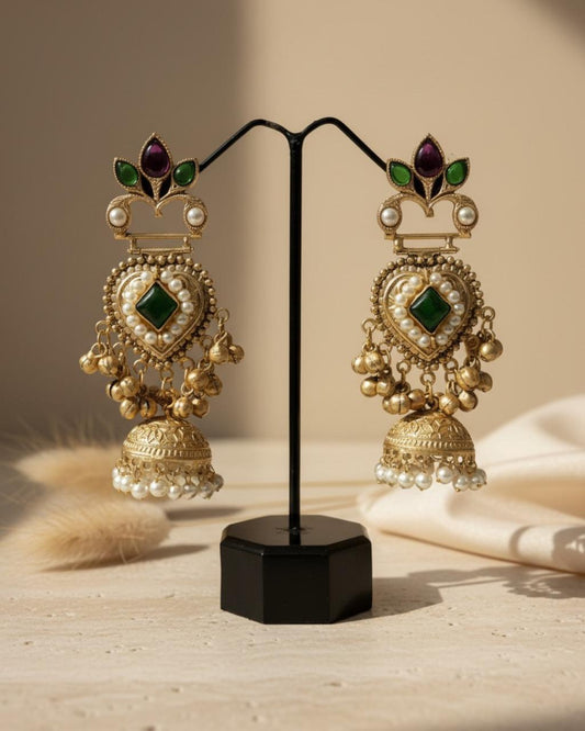 ChicShukra gold‑tone jhumka earrings with purple gem crown, green leaf accents, pearl‑lined heart center, and pearl‑trimmed jhumka bells on a display stand.