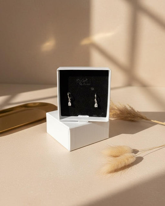 White jewellery box with 'ChicShukra' logo open to reveal a pair of silver‑finish brass teardrop earrings on black velvet.
