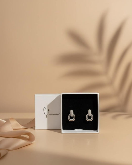 White gift box with 'ChicShukra' branding opened to reveal a pair of silver‑tone brass oval stud earrings on black velvet inside, next to a beige ribbon.
