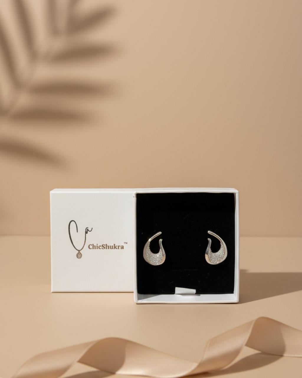 White jewellery box with 'ChicShukra' logo displaying a pair of silver‑tone brass crescent earrings with micro‑pave stones on black velvet insert.