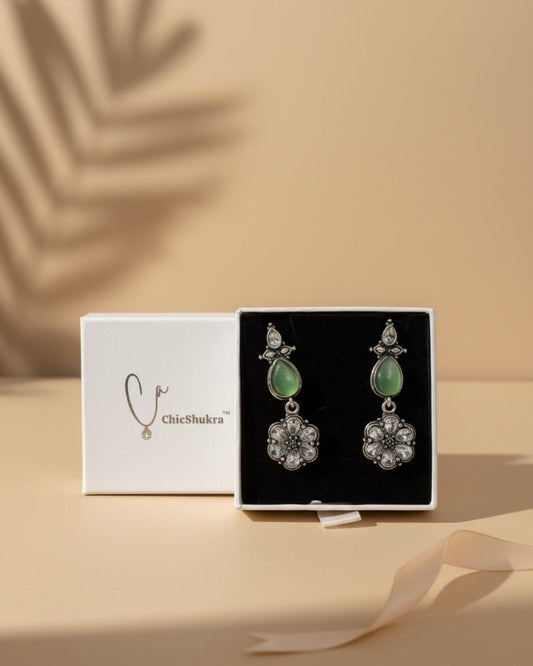 Pair of silver-toned brass earrings with green teardrop stones and crystal floral pendants in a white ChicShukra box on a beige background.