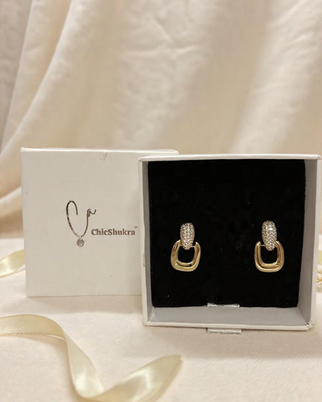 Pair of gold-toned brass ChicShukra earrings with zirconia studs in a white jewellery box with black velvet insert.