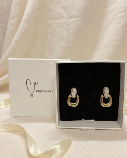 Pair of gold-toned brass ChicShukra earrings with zirconia studs in a white jewellery box with black velvet insert.