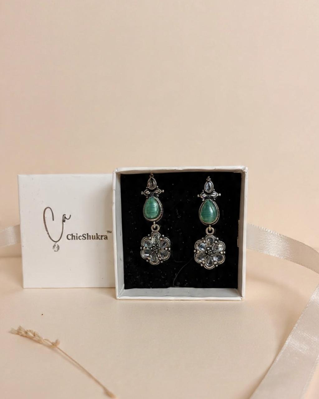 Pair of green stone and silver floral drop earrings by ChicShukra in a white gift box on a beige background.