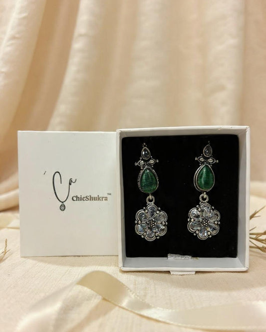 A pair of silver-tone ChicShukra earrings with green pear-shaped stones and flower-shaped clear zirconia pendants displayed in a white jewellery box.