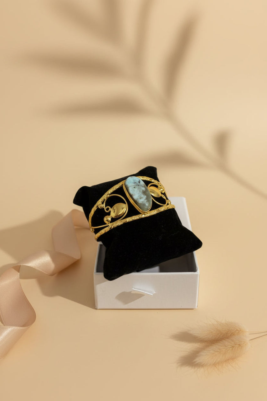 Golden brass cuff bangle with a large oval larimar stone and filigree design from ChicShukra, displayed on a black velvet stand.