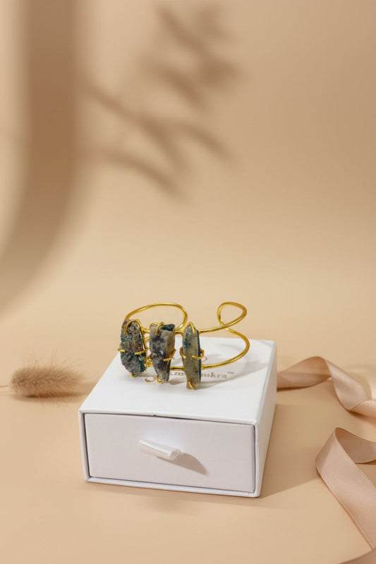 A gold-toned brass bangle with three wire-wrapped Labradorite crystal slabs displayed on a black velvet cushion inside a white jewellery box.