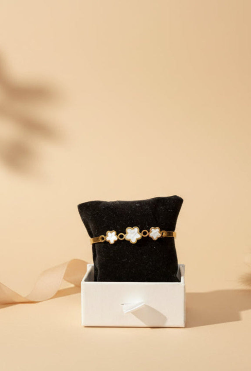 A gold-toned brass bracelet with four white four-petal flower charms displayed on a black jewelry pillow inside a white box