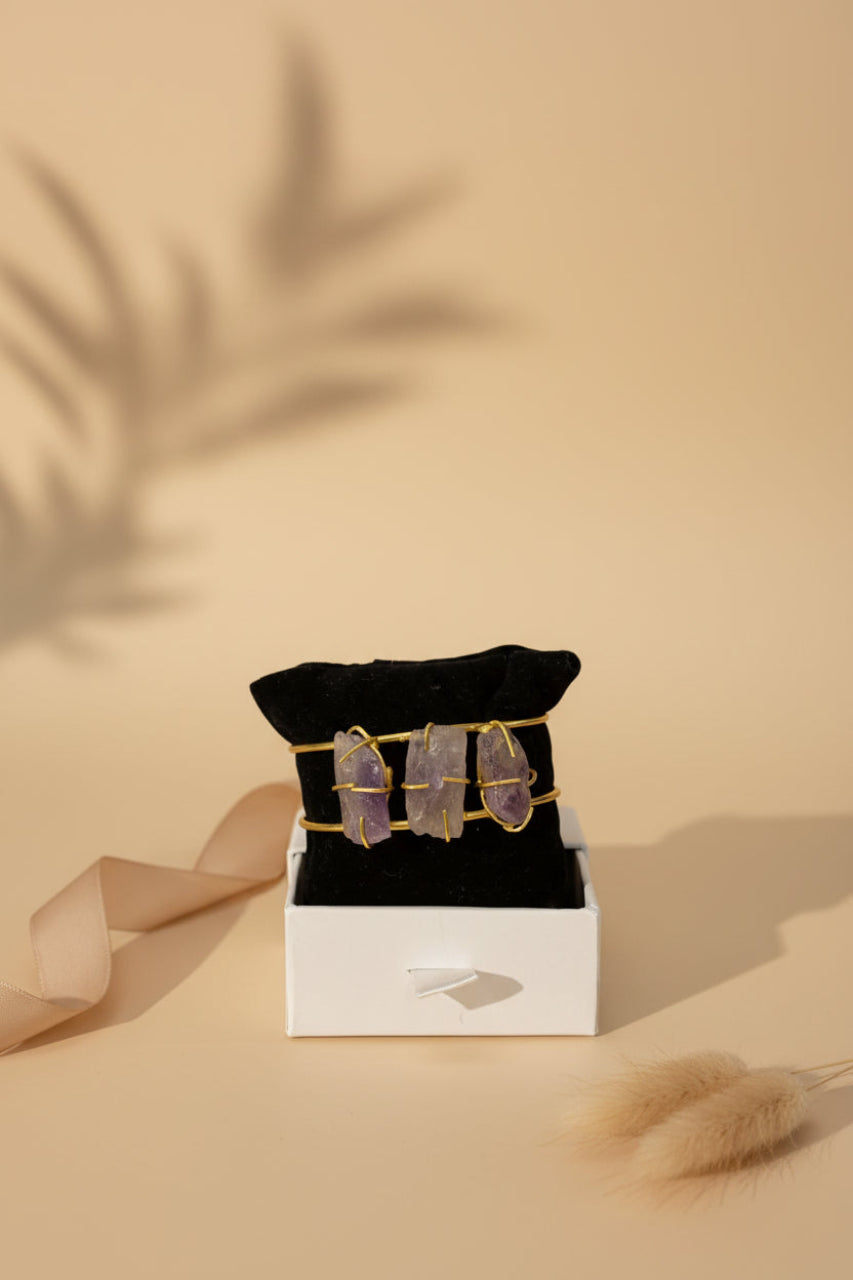 A gold-toned brass bangle with three wire-wrapped amethyst crystal stones displayed on a black velvet stand.