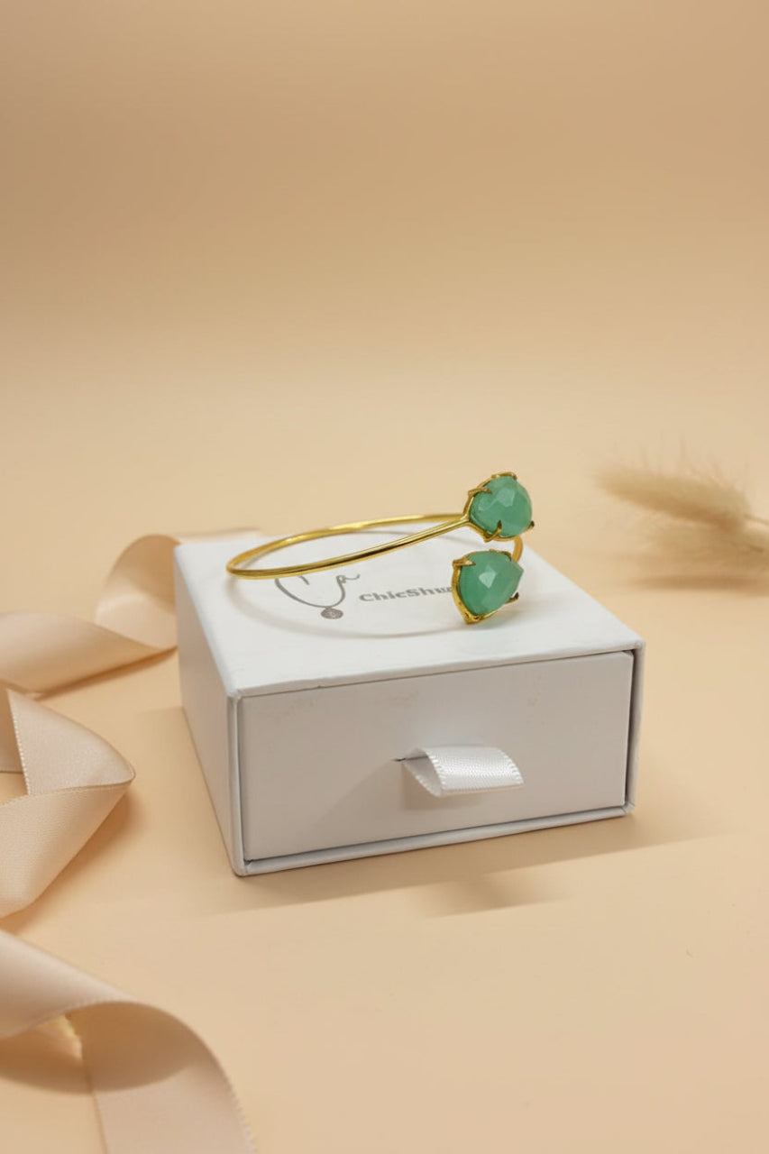 Gold bracelet with green heart-shaped stones on a white box against a beige background