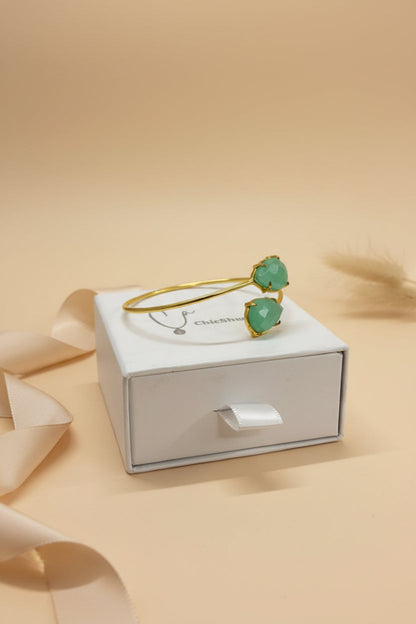 Gold bracelet with green heart-shaped stones on a white box against a beige background