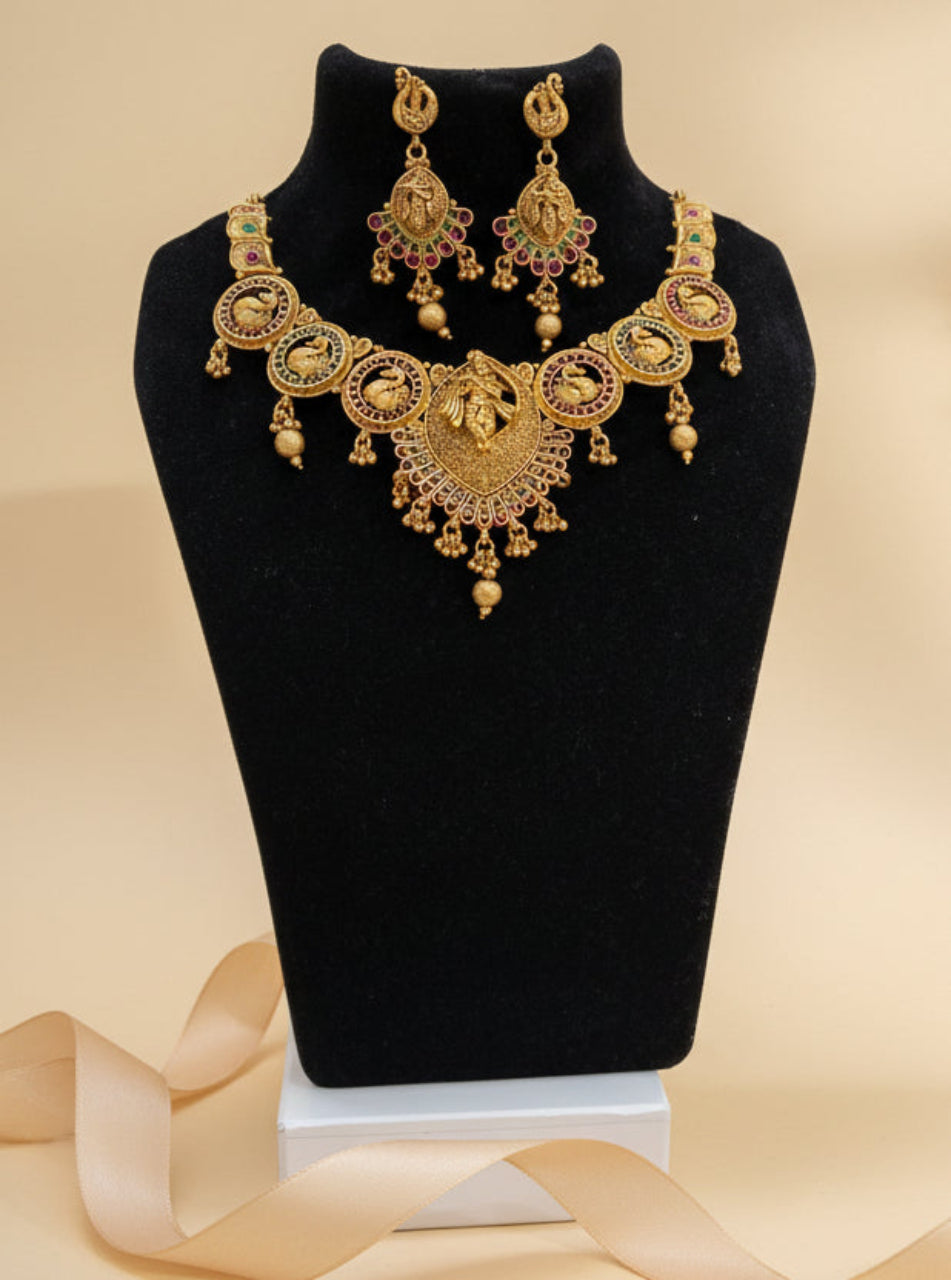 Gold‑plated Chicshukra necklace set with goddess Lakshmi centrepiece, peacock motifs, pink & green stones, and matching earrings on black display.