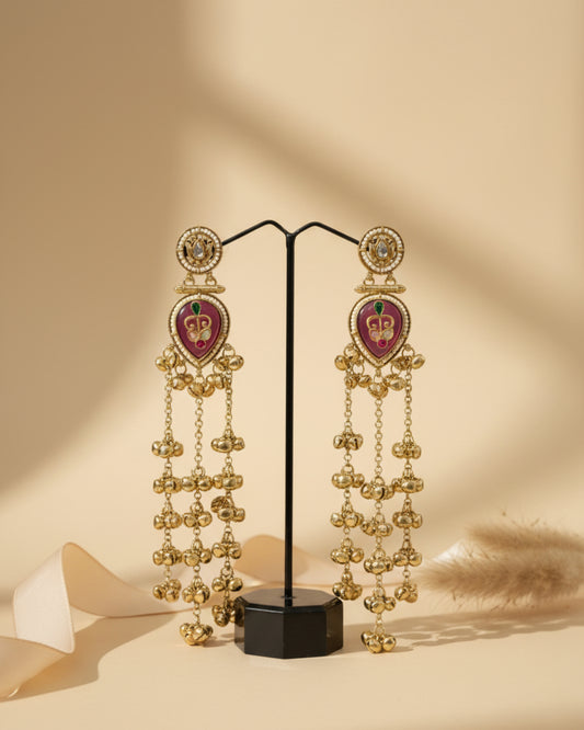 Pair of long gold-plated Kashmiri brass Dehjoor jhumka earrings with emerald green teardrop stones and ghungroo tassels on a stand.