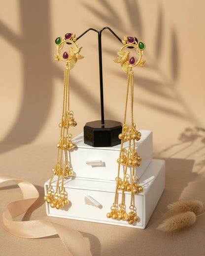Pair of long gold-plated Kashmiri brass Dehjoor jhumka earrings featuring emerald green teardrop stones and cascading ghungroo bell tassels.
