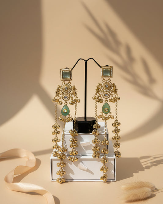 Pair of long gold-plated Kashmiri brass Dehjoor earrings featuring square sea-green stone studs, ornate floral filigree, and cascading ghungroo bell tassels.