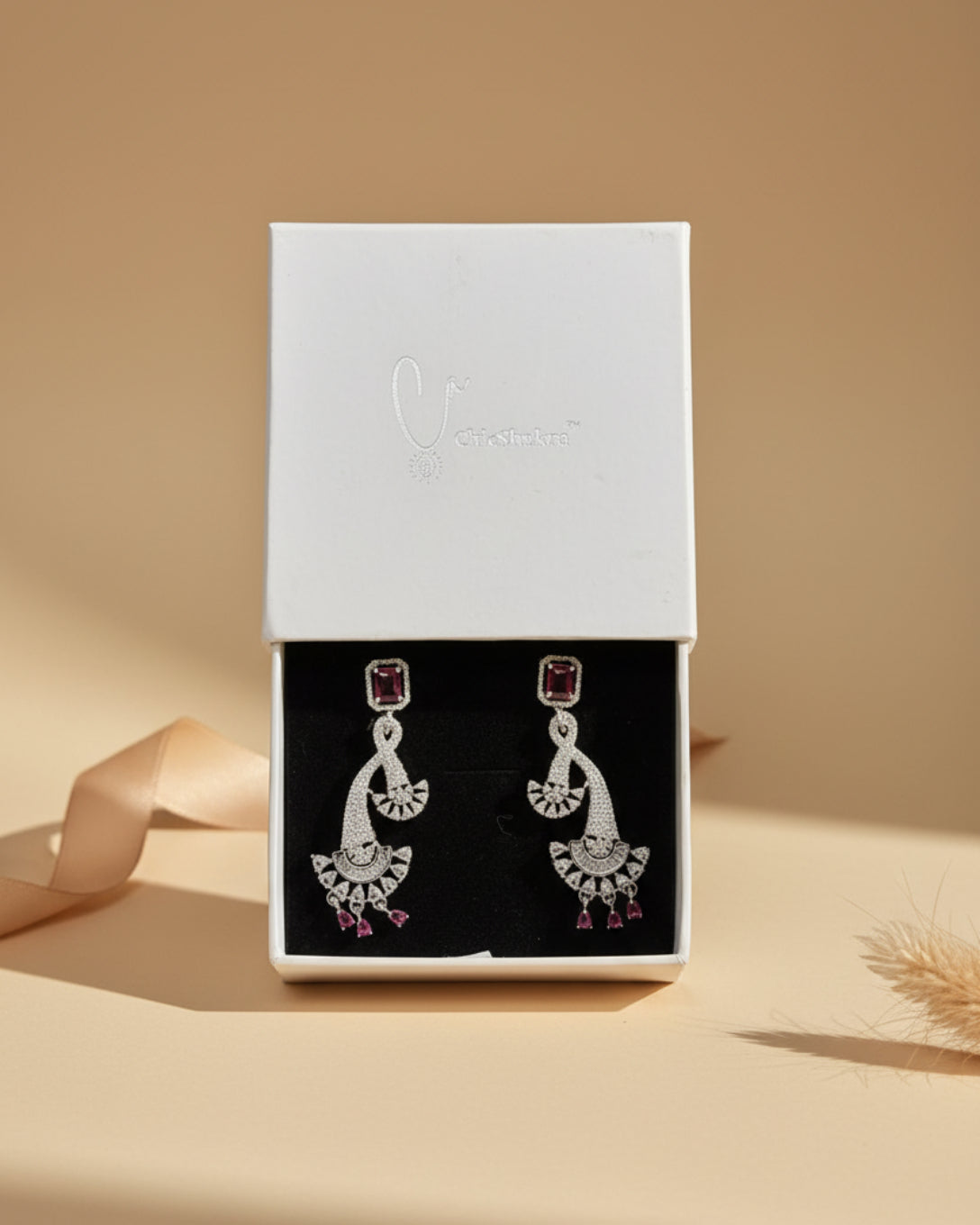 ChicShukra silver-finish AD dangle earrings featuring a ruby red emerald-cut stud and a pave-encrusted fan-shaped drop with ruby accents.