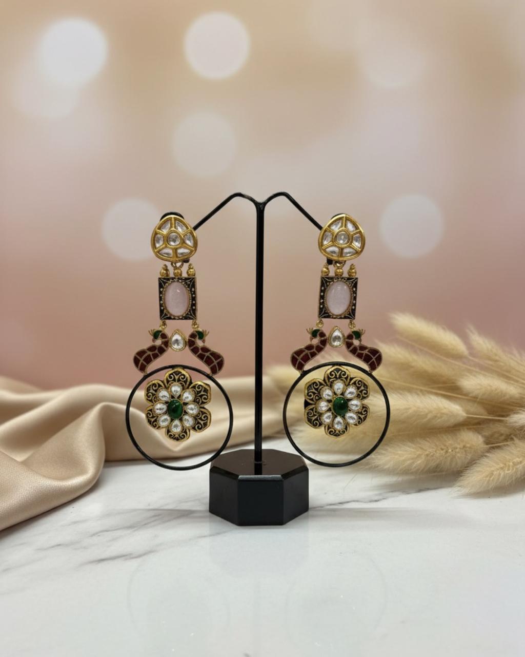 White jewellery box with ChicShukra logo containing gold‑tone brass Kundan earrings with red enamel, green gem and pearl accents on black velvet, next to beige fabric and dried pampas grass.